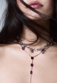 Viacvrstvový strieborný náhrdelník s retiazkami, ozdobený fialovými a priesvitnými drahokamami nepravidelných tvarov a kvapkovým príveskom v tvare písmena Y. Hladká metalická textúra.