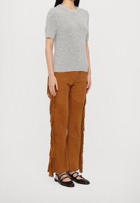 Haut gris en tricot à manches courtes associé à un pantalon en daim marron avec des accents de franges. Des chaussures plates noires complètent la tenue.