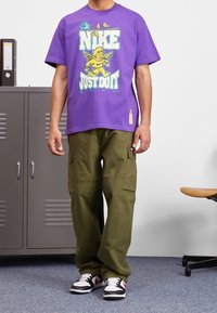 Fialové bavlněné tričko s grafickým potiskem cheruba a nápisem "NIKE JUST DO IT". Kombinováno s olivově zelenými cargo kalhotami a černými/červenými teniskami.