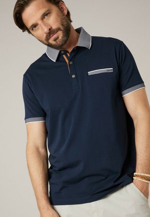 Homme portant un polo bleu marine à manches courtes avec col contrastant, bordure de manchette, détail de poche, et pantalon beige, main dans la poche.