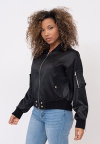Zwarte satijnen bomberjack met rits aan de voorkant, geribbelde manchetten en zijkleppen. Bevat borduurwerk op de borst en een ritszak op de mouw.
