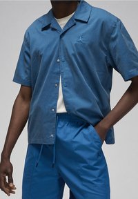 Uomo che indossa una camicia a maniche corte azzurra con bottoni sopra una maglietta bianca e pantaloni blu con stringa, con una mano in tasca su uno sfondo grigio.