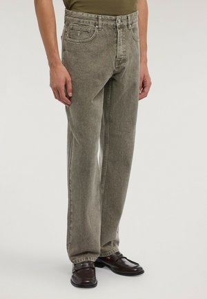Man som bär löst sittande blekta grå jeans och mörkbruna läderloafers, stående mot en enfärgad bakgrund.