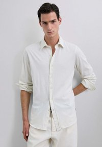 Camisa blanca de manga larga y botonadura, confeccionada en una tela suave, con un corte relajado y mangas remangadas. Diseño simple, sin patrones ni acentos.