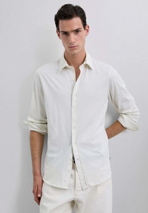 Camisa blanca de manga larga y botonadura, confeccionada en una tela suave, con un corte relajado y mangas remangadas. Diseño simple, sin patrones ni acentos.