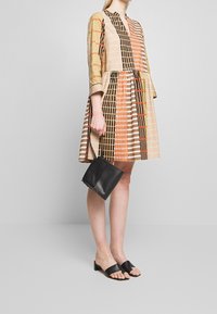 Musterkleid mit langen Ärmeln, das horizontale und vertikale Streifen in Beige, Braun und Orange aufweist. Kombiniert mit einer schwarzen Tasche und Sandalen.