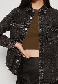 Camicia in jeans neri con una texture sbiadita, caratterizzata da una chiusura con bottoni sul davanti, due tasche sul petto e maniche arrotolate, abbinata a un top corto marrone.