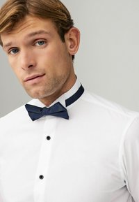 Camisa blanca de vestir con botones negros y una corbata de moño geométrica en color azul marino. El cuello presenta una banda de acento en azul marino a juego.