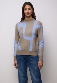 Beiger Pullover mit hellem blauem abstraktem Muster, hohem Kragen, gerippten Bündchen und Saum, kombiniert mit blauen Jeans. Weiche Strickstruktur.