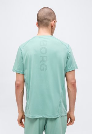 Homme portant un t-shirt de sport à manches courtes vert clair et un short, dos face à la caméra, avec un texte perforé "BORG" sur le t-shirt.