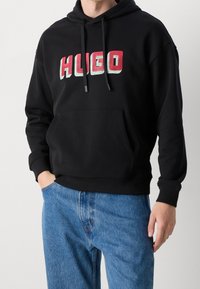 Felpa nera con tasca frontale, caratterizzata dalla scritta "HUGO" in rosso e verde. Vestibilità rilassata con polsini e orlo a coste. Indossata con jeans blu.