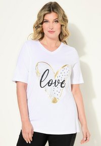 Bílé tričko s krátkým rukávem a V-výstřihem, na kterém je velká zlatá a černá srdcová grafika s nápisem "love" v černé barvě, ozdobená rhinestony.