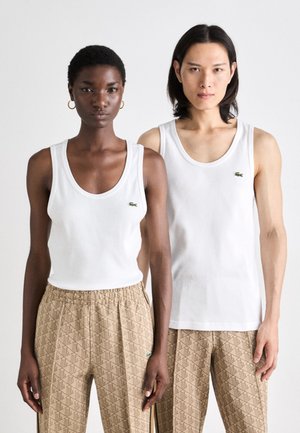 Two individuals wear white, sleeveless tank tops with a green logo. They pair with patterned, beige pants featuring a textured design.