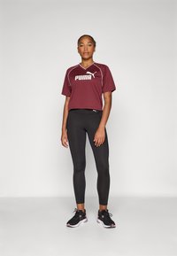Maroon Puma T-shirt with white logo, paired with black leggings and black athletic shoes with pink accents. Model stands against a plain background.