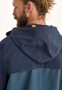 Un homme avec une barbe se détourne, portant une veste à capuche qui est bleu marine sur le haut et turquoise en bas, mettant en valeur des détails de couture.