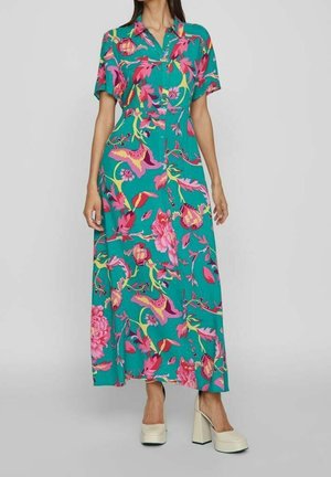 Femme portant une robe maxi à manches courtes boutonnée couleur sarcelle avec de grands motifs floraux roses et jaunes, accompagnée de talons compensés beige.