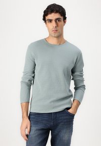 Lichtgroene trui met lange mouwen, een gestructureerd patroon, ronde halslijn en een aansluitende pasvorm, gecombineerd met donkerblauwe jeans.