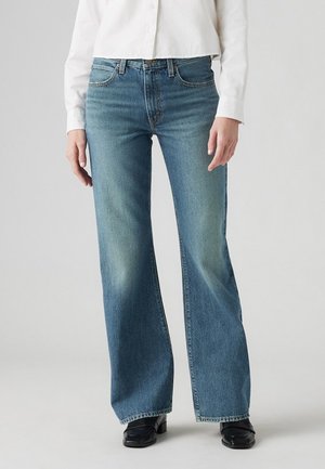 Personne portant un jean bleu à jambes larges, des chaussures noires et une chemise blanche à manches longues boutonnée, debout devant un fond uni.