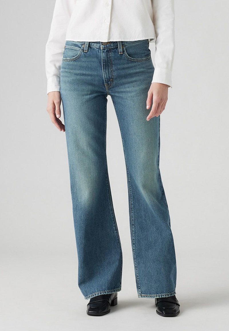 Personne portant un jean bleu à jambes larges, des chaussures noires et une chemise blanche à manches longues boutonnée, debout devant un fond uni.