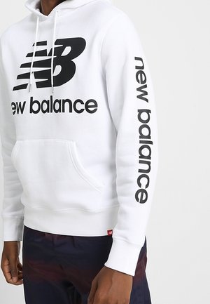 Weißer New Balance Hoodie mit schwarzem Logo und Schriftzug auf der Brust und am rechten Ärmel, getragen von einer Person mit dunklem Hautton.