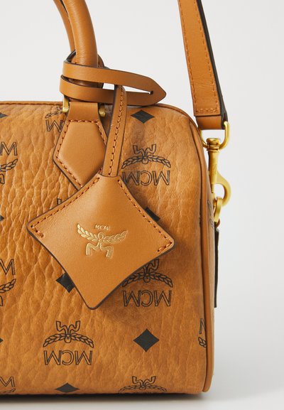 Tan læder MCM håndtaske med sort logo mønster, guldfarvede metaldele, korte hanke og et diamantformet lædermærke fastgjort nær stroppen.