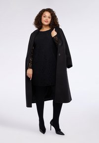 Schwarzer Mantel aus strukturiertem Stoff mit zwei Knöpfen, getragen über einem schwarzen Spitzenkleid. Schwarze Strumpfhosen und spitze Absätze vervollständigen den Look.