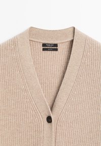 Cardigan en laine beige côtelée avec col en V, un bouton noir unique et étiquette Massimo Dutti à l'intérieur du col.