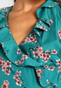 Türaublaue Bluse mit floralem Muster in Pink und Weiß. Verfügt über einen Rüschenausschnitt und eine weiche Stoffstruktur. Nahaufnahme der Designdetails.