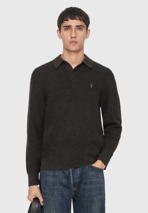 Ung mand iført en mørkebrun polotrøje med lange ærmer og blå jeans, der holder en sort taske i den ene hånd, mod en ensfarvet baggrund.
