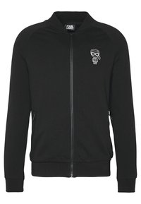 Felpa nera con zip, dotata di colletto e polsini a costine. Presenta tasche laterali e una grafica di un cartone animato bianca sul lato sinistro del petto. Tessuto morbido.