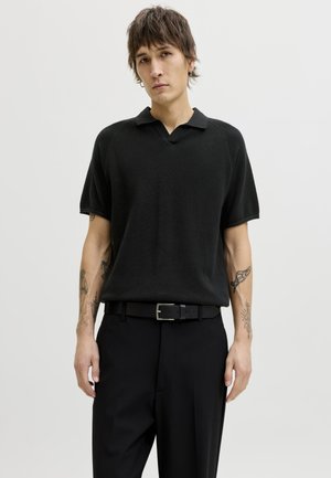 Νεαρό άτομο με κοντά μαλλιά και τατουάζ φοράει μαύρο κοντομάνικο πουκάμισο με υφή, μαύρη ζώνη και μαύρο παντελόνι σε απλό φόντο.