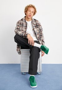 Zöld velúr tornacipők, fehér gumitalp, fekete laza nadrág, fehér póló és kockás dzseki narancs és barna részletekkel.