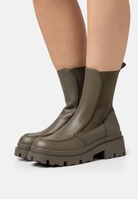 Botas de tobillo verde oliva con un acabado de cuero elegante, paneles laterales elásticos, suelas de plataforma de goma gruesa y un diseño de punta redonda.