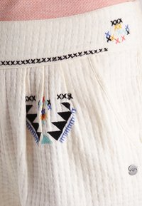 Tissu blanc texturé avec des motifs brodés vibrants en bleu, noir, rouge et vert. Couture décorative le long de la ceinture.