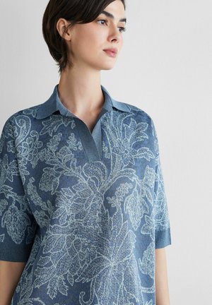 Ung kvinde med kort mørkt hår iført en blå blomsterprintet strikket poloshirt med krave og korte ærmer, der kigger til siden.