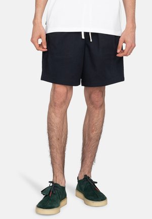 Pantalones cortos azul marino hechos de una tela suave, con dos bolsillos delanteros y una cintura con cordón. Combinados con zapatos de ante verdes.