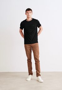 Schwarzes T-Shirt mit kurzen Ärmeln und Rundhalsausschnitt, kombiniert mit hellbraunen Slim-Fit-Hosen und weißen Sneakers. Einfaches Design und neutrale Farben.