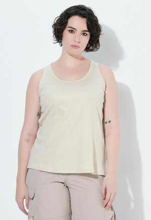 Persona joven con cabello corto, rizado y oscuro, lleva una camiseta sin mangas beige y pantalones cargo claros, de pie frente a un fondo blanco liso.