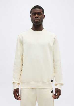 Mladý muž oblečený v krémově světlé mikině a odpovídajících kalhotách, stojící čelem k divákovi před jednobarevným světlým pozadím.