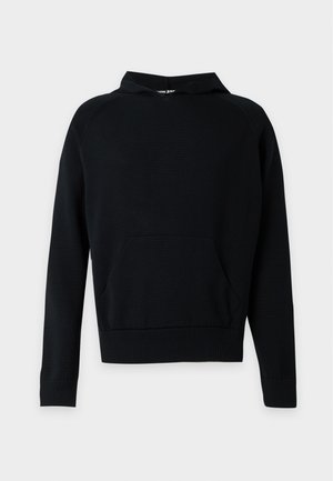 Homme portant un sweat à capuche noir avec poche avant, pantalon beige à jambes larges avec des détails de boutons aux ourlets, et des chaussures noires, debout devant un fond uni.