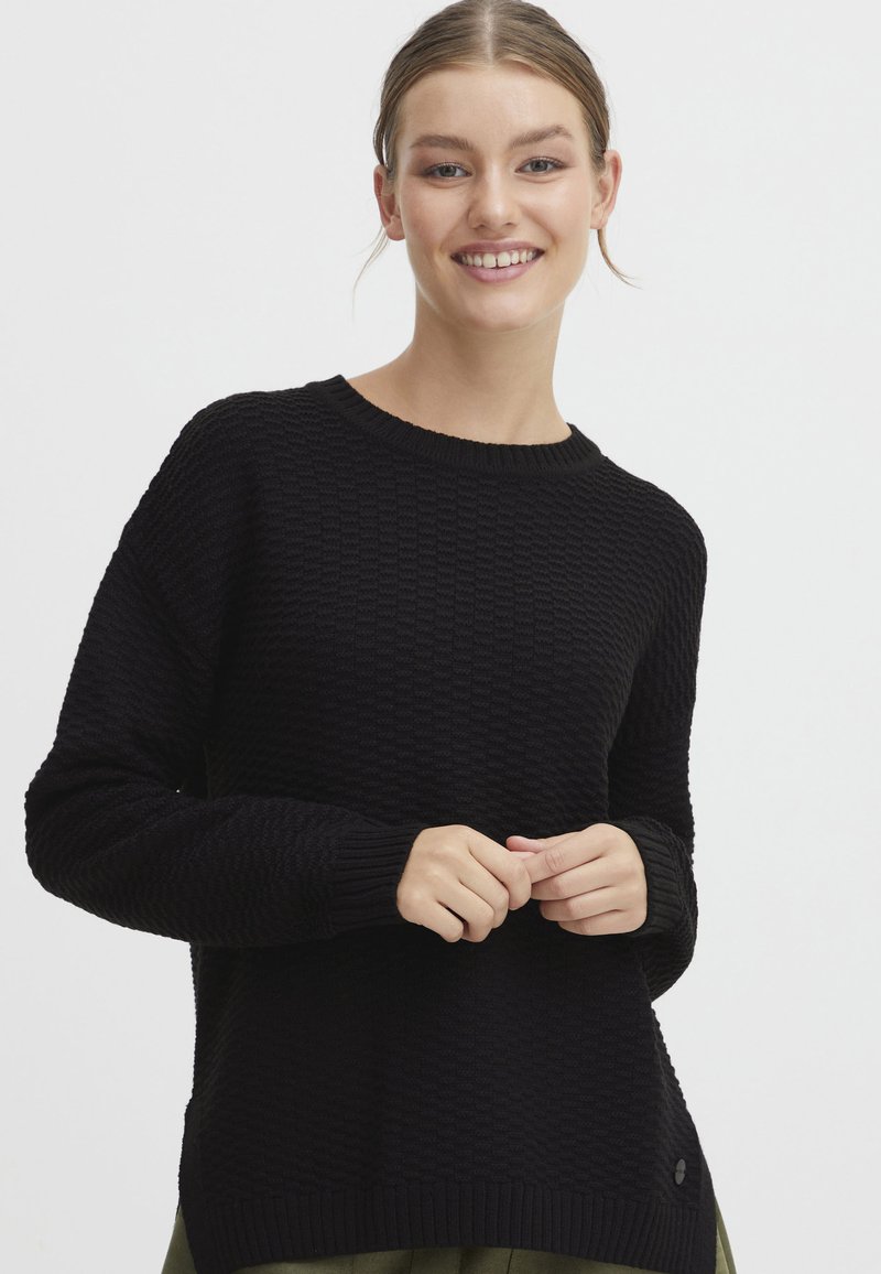 Schwarzer Strickpullover mit strukturiertem Muster, runder Halslinie und gerippten Bündchen. Bemerkenswertes seitliches Detail ist ein kleines Logo-Element.