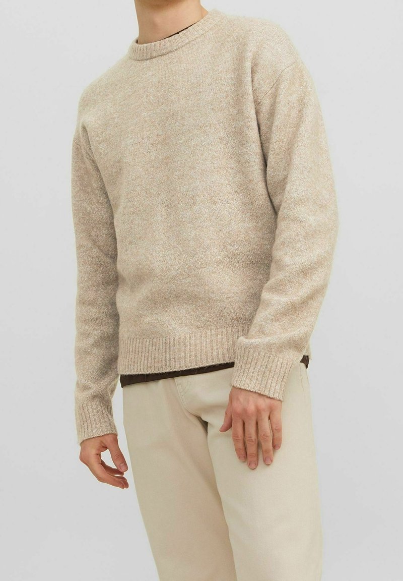 Beiger Strickpullover mit Rundhalsausschnitt, gerippten Bündchen und Saum. Weiche Textur, leicht meliertes Muster. Über einem dunklen Oberteil getragen.