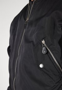 Schwarze Bomberjacke mit Reißverschluss, zwei seitlichen Taschen mit Zippern und einem Druckknopfdetail an der Taschenklappe. Glatte Materialtextur.