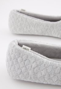 Graue Hausschuhe aus Stoff mit strukturiertem Muster, weichem Innenfutter und einer abgesteppten Kante. An der Seite befindet sich ein kleines Etikett mit der Aufschrift „NEXT“.