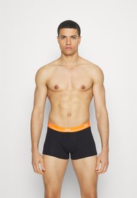 Black boxer shorts with an orange logo waistband. The fabric appears smooth and stretchy. Model stands in a neutral pose against a light background.