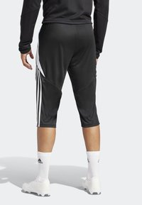 Pantaloni sportivi neri con un design a sigaretta, dotati di strisce laterali bianche e una texture liscia. Abbinati a calzini bianchi al polpaccio e scarpe da calcio.