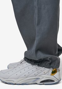 Hvide lædersneakers med perforerede detaljer og metalliske guld accenter. Båret med grå bukser med opretstående kant, der hænger over skoene.