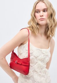 Νεαρή γυναίκα που φοράει λευκό φόρεμα με λουλούδια κρατά μικρή κόκκινη δερμάτινη τσάντα ώμου, κοιτάζοντας προς το πλάι σε απλό φόντο.