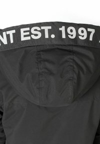 Svart huva jacka med en jämn textur, med ett framträdande vitt band vid kragen som har texten "EST. 1997." och en ren, minimalistisk design.