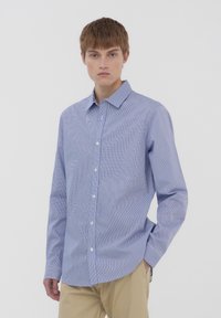 Camicia azzurro chiaro con sottili strisce bianche, colletto classico e maniche lunghe, realizzata in un tessuto liscio. Indossata con pantaloni beige.